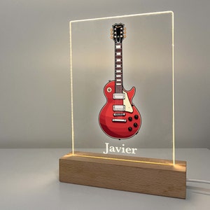 May include: A red electric guitar illustration on a clear acrylic panel, illuminated by an LED light. The guitar is detailed with black accents and white pickguard. The name "Javier" is printed below the guitar. The base is made of light-colored wood.