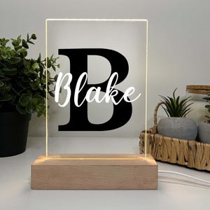 May include: A personalized LED night light with a wooden base. The clear acrylic panel displays a large black letter "B" with the name "Blake" in white script. The light emits a warm glow, perfect for a bedroom or nursery.