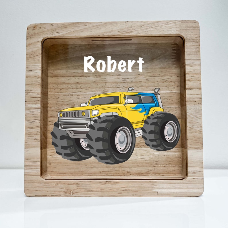 May include: A wooden piggy bank with a slot on top. The front of the bank features a yellow monster truck with blue flames and the name "Robert" printed above it.