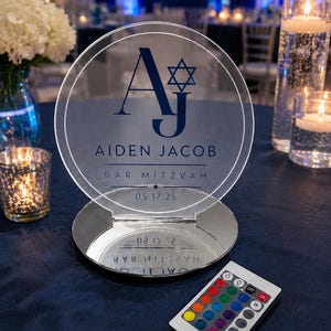 May include: A circular clear acrylic sign with a dark blue monogram AJ, a Star of David, and the text Aiden Jacob Bar Mitzvah 05.17.25. The sign sits on a round reflective base and includes a small white remote control with multi-colored buttons.