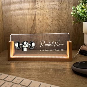 May include: A wooden desk nameplate with a clear acrylic front. The nameplate features a dumbbell graphic and the text "Rachel Kim, Personal Trainer." The base and edges of the nameplate are illuminated with warm white light.