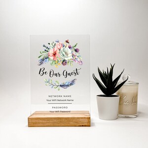 May include: A clear acrylic sign with a floral design and the text "Be Our Guest". The sign has a wooden base and includes spaces for the WiFi network name and password.