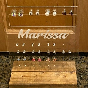 May include: A clear acrylic earring holder with three rows of earring posts. The holder has a wooden base and the name "Marissa" is printed on the front.