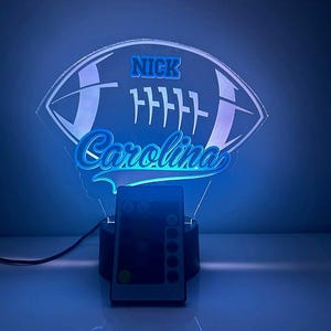 May include: A blue and white football-shaped night light with the name "Nick Carolina" on it. The light is on a black base with a remote control.