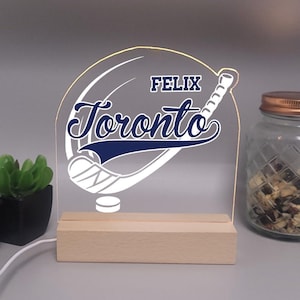 May include: A clear acrylic LED night light with the name "Felix" above the word "Toronto" in a stylized font. The design includes a hockey stick and a hockey puck. The light sits on a wooden base.
