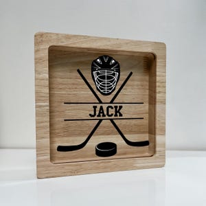 May include: A wooden shadow box with a hockey theme. The design includes a hockey helmet, crossed hockey sticks, a puck, and the name "JACK" in a bold font. The box has a natural wood grain finish.