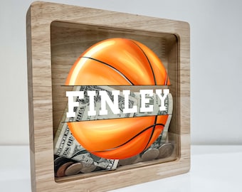 Puede incluir: Una caja de madera para guardar dinero con un diseño de baloncesto y el nombre "FINLEY" impreso en la parte delantera. La caja está llena de monedas y billetes.