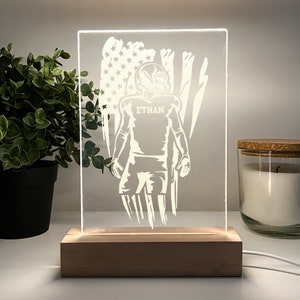 Könnte beinhalten: Ein weißes Acryl-Nachtlicht mit Holzsockel. Das Licht zeigt die Silhouette eines Footballspielers vor einer amerikanischen Flagge. Das Trikot des Spielers trägt den Namen "ETHAN" aufgedruckt.