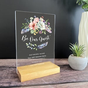 May include: A clear acrylic sign with a floral design and the text "Be Our Guest". The sign also includes the text "Network Name" and "Password" with spaces for you to write in your own information. The sign is on a wooden stand.