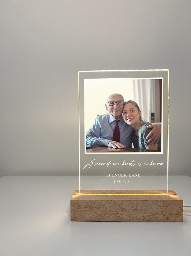 May include: A white acrylic light-up photo frame with a picture of an older man and a woman smiling. The text "A piece of our hearts is in heaven" is written in cursive on the frame. Below the picture, the text "SPENCER LANE 1946-2019" is written in white.