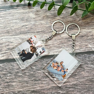 Könnte beinhalten: Zwei transparente Acryl-Schlüsselanhänger mit silbernen Ketten. Ein Schlüsselanhänger zeigt ein Foto einer Frau und eines Kindes, der andere zeigt ein Foto einer Frau und mehrerer Kinder. Der Text "Blessed" ist auf der Unterseite des Schlüsselanhängers mit dem Gruppenfoto aufgedruckt.