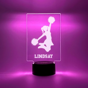 May include: A white silhouette of a cheerleader with pom-poms, jumping in the air. The name "LINDSAY" is written below the silhouette. The image is on a clear acrylic plaque with a black base.