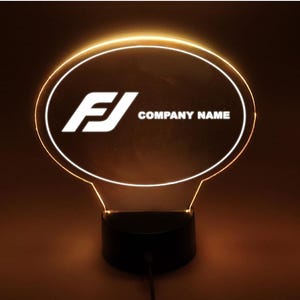 May include: An illuminated LED lamp with a circular design. The lamp displays a white logo and the text "COMPANY NAME" within an oval shape. The base is black, and the light emits a warm glow. The lamp is powered by a cord.