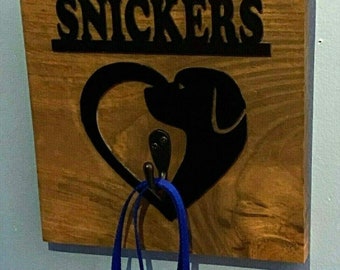 Puede incluir: Gancho de madera para la correa de un perro con una silueta negra de un perro en forma de corazón. El gancho está etiquetado como "SNICKERS" y tiene una correa azul colgando de él.