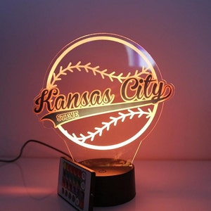 May include: A baseball-themed night light with the words "Kansas City" in a stylized font. The name "Steve" is below the city name. The light has a black base and a remote control. The light is glowing orange.