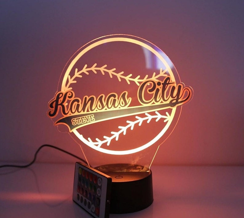 May include: A baseball-themed night light with the words "Kansas City" in a stylized font. The name "Steve" is below the city name. The light has a black base and a remote control. The light is glowing orange.
