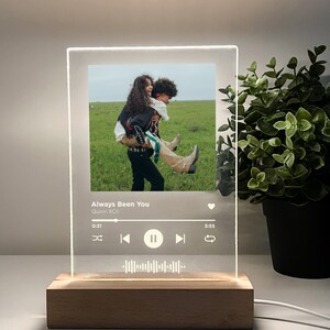 Op de afbeelding: Een helder acryl fotolijst met een houten basis. De lijst toont een foto van twee mensen in een veld, met een muziekspelerinterface onder de foto. De muziekspelerinterface toont de titel van het nummer "Always Been You" van Quinn XCII, de duur van het nummer en de afspeelbesturing.