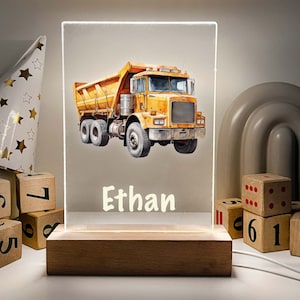 Könnte beinhalten: Ein gelber Spielzeug-Kipplaster mit einer weißen Basis und dem Namen "Ethan" in weiß auf der Basis. Der Laster steht auf einer Holzbasis mit einem Licht dahinter.
