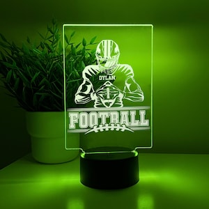 Op de afbeelding: Een wit silhouet van een American footballspeler die een voetbal vasthoudt met de naam "DYLAN" en het woord "FOOTBALL" eronder. De afbeelding is op een heldere acrylbasis met een zwarte basis.