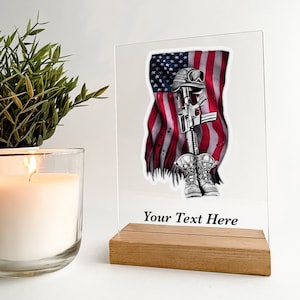 May include: A clear acrylic plaque with a black and white illustration of a folded American flag, a rifle, a military helmet, and boots. The text "Your Text Here" is printed below the illustration. The plaque is on a wooden stand.