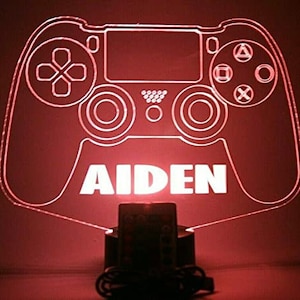 May include: A white, translucent, illuminated PlayStation controller with the name "AIDEN" printed below. The controller is lit with a red light and is sitting on a black base.