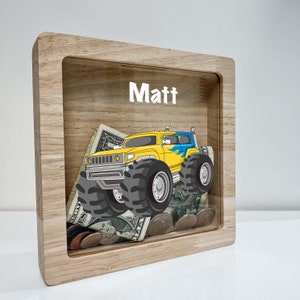 May include: A wooden money box with a clear front panel. The box has a yellow monster truck graphic and the name "Matt" written on the top. The box is filled with coins and bills.