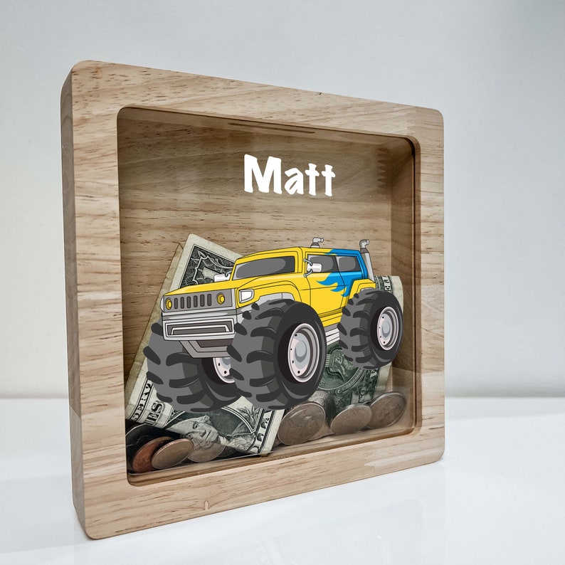 May include: A wooden money box with a clear front panel. The box has a yellow monster truck graphic and the name "Matt" written on the top. The box is filled with coins and bills.