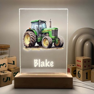 Op de afbeelding: Een gepersonaliseerd nachtlampje met een illustratie van een groene tractor. De tractor staat tegen een zachte achtergrond, met de naam "Blake" in het wit verlicht. Het licht staat op een houten voet.