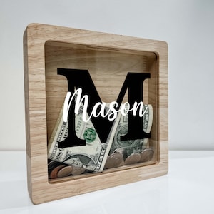 May include: Wooden money box with a clear front panel. The front panel has a black letter "M" with the name "Mason" written in white cursive script. There are coins and dollar bills inside the box.