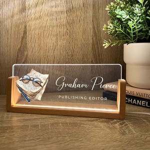 May include: A wooden desk nameplate illuminated from within, displaying the name "Graham Pierce" and the title "Publishing Editor". The design features a stack of old paper, spectacles, and a pen.