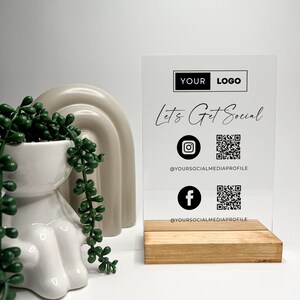 May include: A clear acrylic sign with a wooden base, featuring the text "Let's Get Social" with Instagram and Facebook icons and QR codes. The sign also includes the text "YOUR LOGO" and "@YOURSOCIALMEDIAPROFILE".