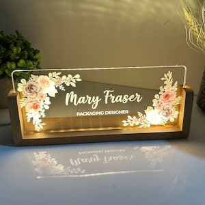 May include: A wooden sign with a clear acrylic insert featuring a floral design and the text "Mary Fraser Packaging Designer". The sign is illuminated from behind.