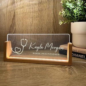 May include: A wooden desk nameplate with a clear acrylic front. The nameplate is illuminated with a warm light and features the name "Kayla Morgan" and the title "Nurse Practitioner" along with a stethoscope graphic.