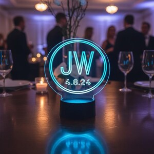 May include: A circular acrylic sign with a glowing cyan neon-style border, featuring the letters JW and the date 4.8.24 in the center. The sign is mounted on a solid black circular base and emits a bright blue light onto the table surface.