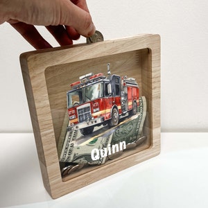 May include: A wooden piggy bank with a clear front panel. The front panel features a red fire truck with the name "Quinn" printed below. The piggy bank is filled with coins and bills.