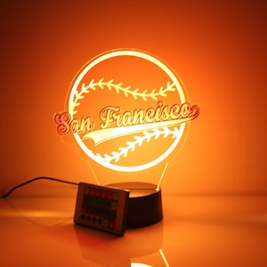 Puede incluir: Una luz nocturna 3D de acrílico blanco con forma de pelota de béisbol con el texto "San Francisco" en una fuente blanca de escritura. La luz nocturna está iluminada con un brillo amarillo cálido y tiene una base negra con un control remoto.