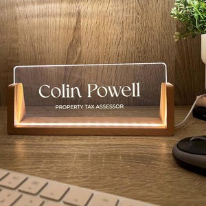 May include: A lighted desk nameplate with the name "Colin Powell" and the title "PROPERTY TAX ASSESSOR" in white lettering. The nameplate is made of clear acrylic and sits in a wooden base. The base has a warm, glowing light.