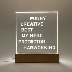 Może przedstawiać: Biały znak akrylowy z napisami "FUNNY CREATIVE BEST MY HERO PROTECTOR HARDWORKING" ułożonymi pionowo. Znak jest podświetlany od tyłu i stoi na drewnianej podstawie.