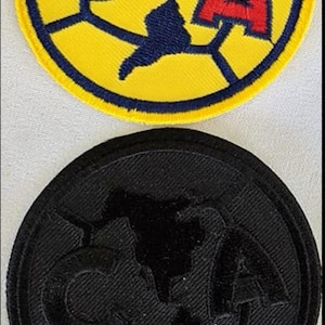 Puede incluir: Dos parches bordados, uno amarillo con un logotipo azul y rojo, y otro negro con un logotipo negro. Los logotipos son una C y una A estilizadas con un globo en el medio.