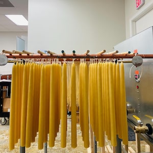 Peut inclure: Une grande collection de bougies effilées jaunes suspendues à des tiges en bois sur un support en cuivre. Les bougies sont uniformes en forme et en couleur, suspendues par leurs mèches dans un atelier avec une machine métallique visible.