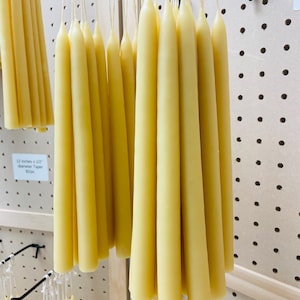 May include: A collection of pale yellow taper candles hanging from a display rack. Each candle features a smooth, cylindrical body that tapers to a point at the top, with a white wick extending from the center. A label indicates 12 inches by 0.5 inches.