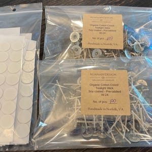 May include: Clear plastic bags containing candle-making supplies. The bags hold white wicks, metal wick tabs, and adhesive dots. The labels read "Organic Cotton Creed Tealight Wick" and "Soy coated - Pre-tabbed W-24".