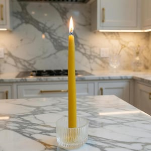 May include: A lit yellow candle in a clear, ribbed glass candle holder. The candle is tall and slender, with a bright flame. The candle holder sits on a marble countertop in a kitchen setting.