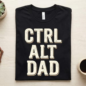Può includere: Una t-shirt nera a maniche corte con la scritta "CTRL ALT DAD" stampata in un grande carattere bianco sporco dall'aspetto usurato. Le lettere presentano un sottile effetto ombra e la maglietta è distesa con un'etichetta visibile sul collo.