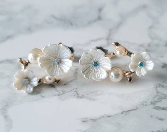 May include: A pair of white flower earrings with gold accents and blue gemstones. The earrings are shaped like a branch with three flowers.