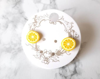 May include: A pair of yellow lemon slice stud earrings on a white card with a decorative border.