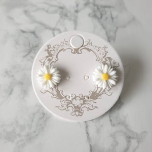 May include: A pair of white daisy stud earrings with yellow centers on a white card with a decorative design.
