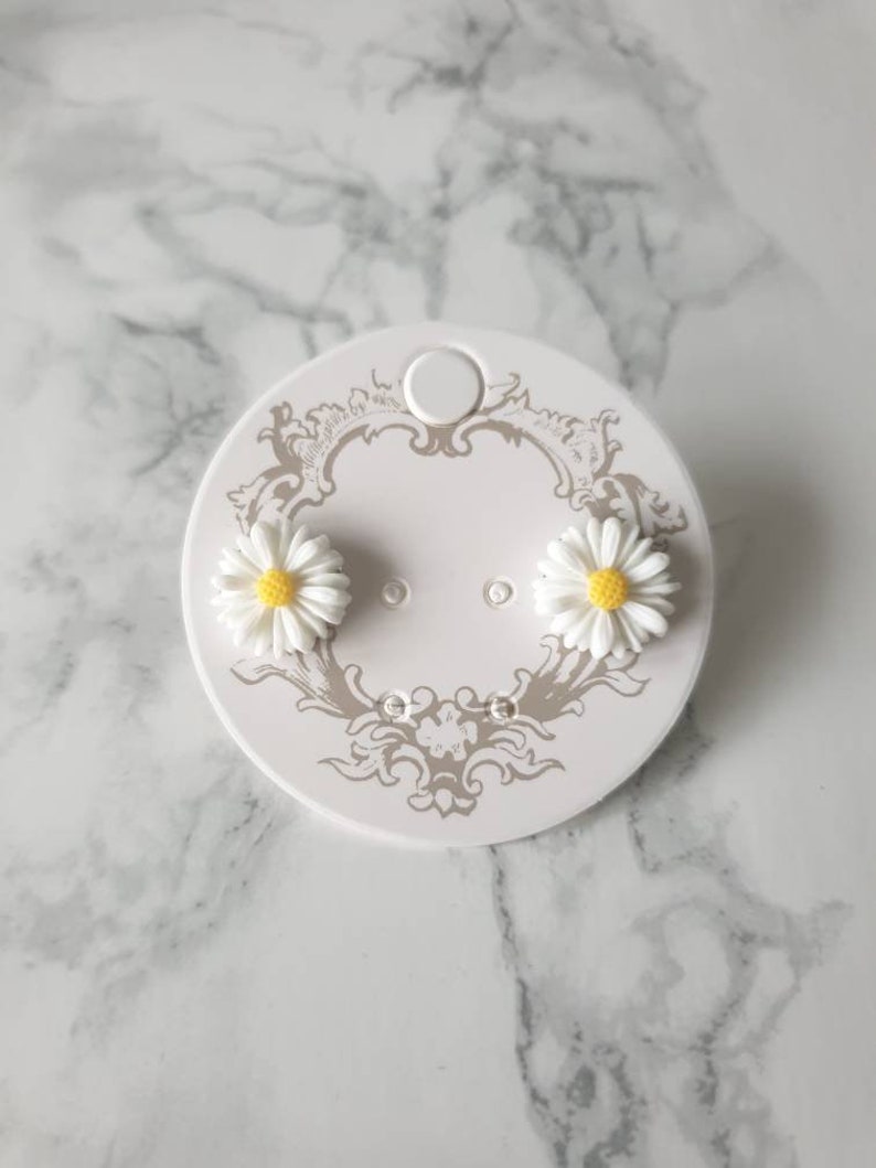 May include: A pair of white daisy stud earrings with yellow centers on a white card with a decorative design.