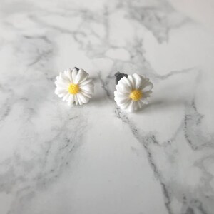 May include: A pair of white daisy earrings with yellow centers. The earrings are on a white marble surface.