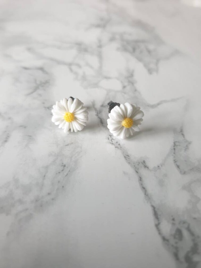 May include: A pair of white daisy earrings with yellow centers. The earrings are on a white marble surface.